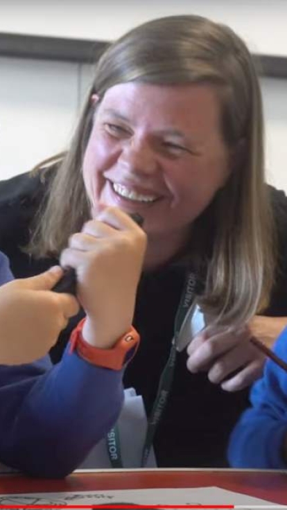 Three children in blue school uniforms are seated at a table, engaged in a creative activity. The child on the left, wearing glasses, holds a black object, possibly a pen or marker. The child in the middle, with long hair, is leaning forward and partially obscured. The child on the right is drawing on paper with a pencil or crayon.