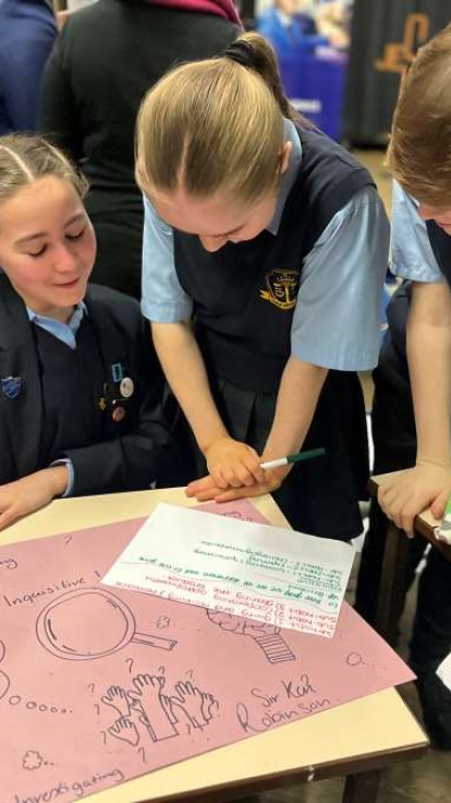 A group of students in school uniforms are gathered around a table working on a collaborative project. On the table is a large pink poster featuring colorful drawings, question marks, raised hands, and words like 'Imagination,' 'Creativity,' and 'Super Kid.' One student holds a printed paper, another writes with a green marker, and another holds an envelope or folder. Other students are visible in the background working at separate tables.
