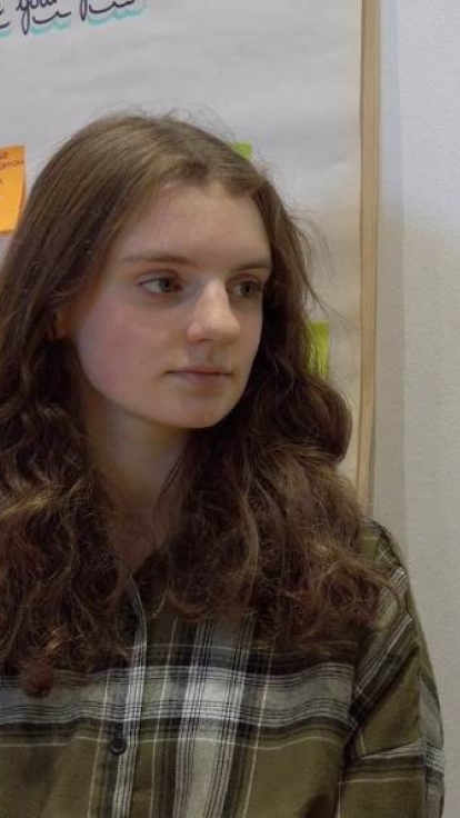 Two individuals with blurred faces stand side by side. The person on the left has long, wavy hair and wears a green plaid shirt. The person on the right has short, curly hair and wears a striped sweater. Behind them is a wall displaying colorful sticky notes and a large sheet of paper with handwritten blue text at the top.