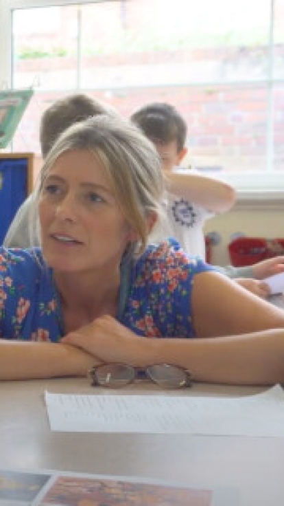 A classroom scene with three students and a teacher seated around a table. The student on the left has long braided hair, a white shirt, and a pink headband. The middle student has brown hair tied back and is wearing a grey sweater, resting their chin on their hand. The teacher on the right has short blonde hair, glasses, and a floral blouse, leaning forward with arms crossed on the table. The background features colorful artwork on the walls, blue storage bins, bookshelves, and windows letting in natural l