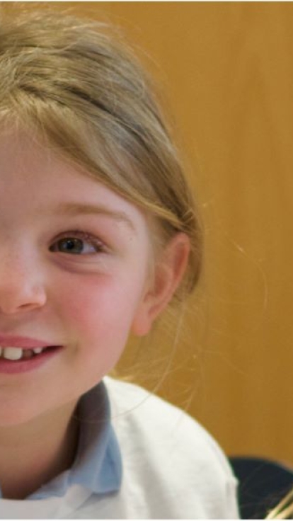 Three children wearing light-colored shirts are sitting closely together, appearing to engage in a conversation or activity. The background is a plain wooden wall.