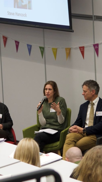 A panel discussion takes place in a lecture hall with five speakers seated at the front, each holding a microphone. The audience, seated in tiered rows, listens attentively and takes notes
