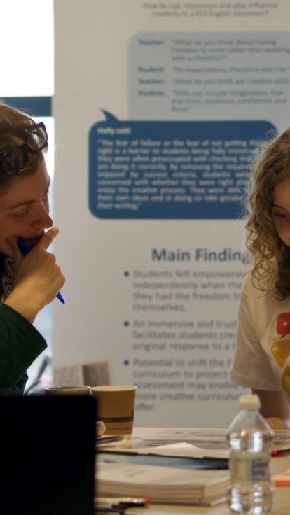 Two people sit at a table engaged in discussion or study. One wears a green sweater and holds a pen; the other wears a white t-shirt with speech bubble graphics. The table is scattered with papers, notebooks, a water bottle, and coffee cups.