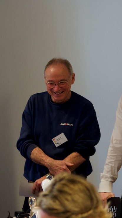 Three people stand in conversation in a room. On the left, a young person wears a white t-shirt with colorful graphics; in the center, an older person wears a dark blue sweater; and on the right, a person wears a white shirt and tie.