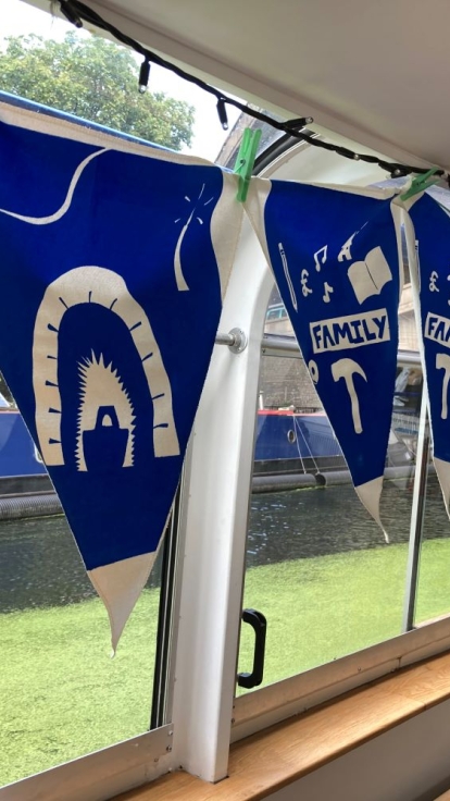 Blue and white triangular flags with words like 'FAMILY' and 'THE SUN,' and images such as a horse, archway, and musical notes, hang inside a boat with windows overlooking water and greenery.