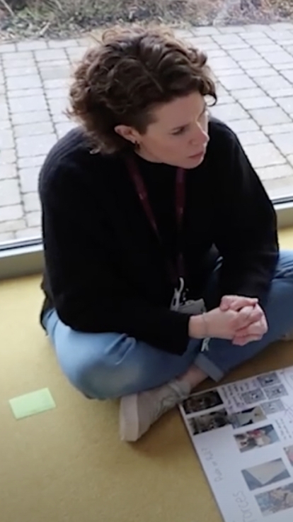 A teacher and three young students sit on the classroom floor, looking at a large poster with images and text. The teacher wears a black sweater and blue jeans; the students are in purple school uniforms. The classroom has yellow walls and a large window revealing an outdoor paved area.