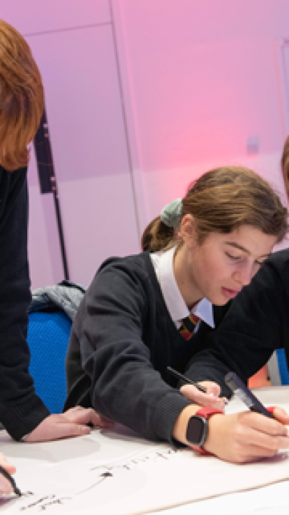 Three students in school uniforms are seated at a table, focused on their work. They are wearing black sweaters with a school crest and ties featuring red, yellow, and black stripes. Their faces are blurred for privacy. In the background, a person is standing and holding papers in a classroom setting