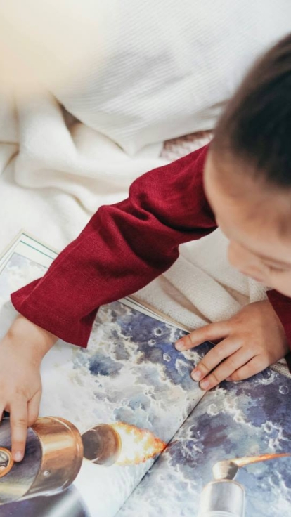 A child in a red shirt sits on a white blanket, pointing at a book illustration of a rocket launching into space. The rocket image shows flames coming from its engines.