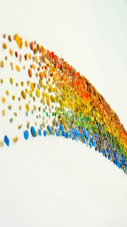 A white wall displays a curved, rainbow-gradient arrangement of small, colorful objects. The gradient flows from red on the left through orange, yellow, green, blue, and ends with purple on the right. The objects vary in shape, including stars, circles, squares, and abstract forms