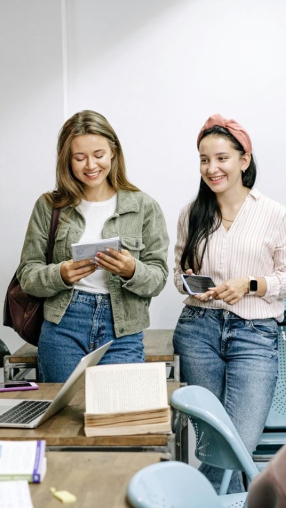 Five people in a classroom. One seated at a desk with a laptop and books, while four others stand nearby, two holding notebooks or papers. Blue chairs are arranged in rows, with a white wall in the background.