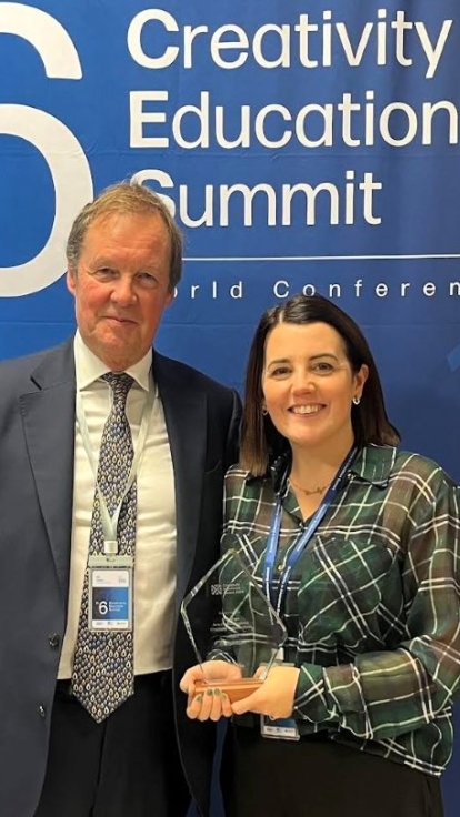 Two individuals with conference badges stand in front of a blue backdrop at the 6th Creativity in Education Summit, featuring OECD and UNESCO logos. One holds an award.