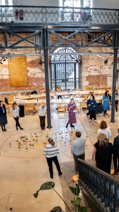Interior of a spacious, industrial-style venue with high ceilings, exposed brick walls, and large windows. People are engaged in activities and conversations around tables and displays. The scene is viewed from a staircase with metal railings in the foreground.