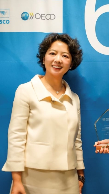 Two individuals stand in front of a blue backdrop featuring logos for the Global Institute of Creative Thinking, UNESCO, and OECD, along with the text '6 Education Summit World Conference.' The person on the left wears a white outfit, and the person on the right wears a green plaid shirt and holds an award.