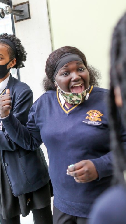 Students in school uniforms stand by a window, with two writing or drawing on it using markers. The word 'VIRUS' is written in red, surrounded by related images and drawings. Modern buildings are visible through the window in the background.