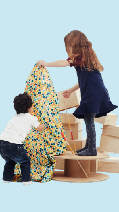 Two children play with a colorful patterned fabric, draping it over a large wooden spool. The child on the left wears a white shirt and jeans; the child on the right wears a navy blue dress and grey leggings. They are surrounded by wooden objects like spools and rectangular blocks, with a pile of plaid fabric on the floor to their right. The background is light blue with a cloud illustration in the top left corner.
