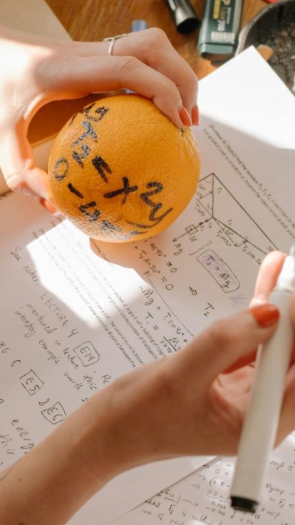 Person holding an orange with the equation 'y = x + 2' written on it, and a white marker in the other hand. On the table are handwritten notes, printed math problems, and a closed book, suggesting a study session focused on functions and derivatives.
