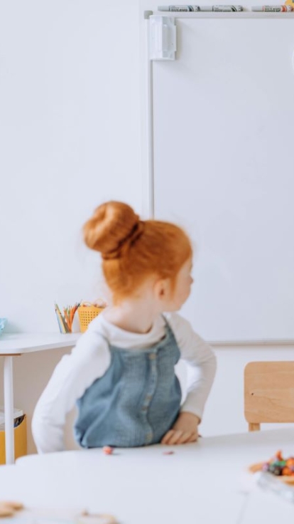 Three children in a classroom: one stands at a whiteboard drawing or writing, while two others sit at a table working with small objects and papers. The whiteboard displays colorful drawings and numbers from 5 to 10.