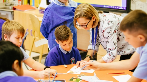An image of a group of young people sat around a school table, using a paintbrush and paper to paint a picture. Their teacher is leaning over the table showing them a technique. 