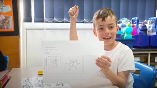 A young boy holds a piece of his work up with a smile on his face. 