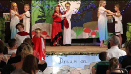 Six young people stand on stage in pairs, dancing together. They are wearing colourful clothing and wearing crowns. 