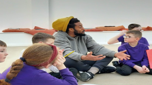 Children in purple jumpers sit on the floor around a man wearing a yellow beanie hat.