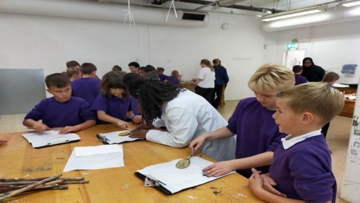 Children wearing purple jumpers and a woman wearing a white coat stand round a table while print painting with household objects.