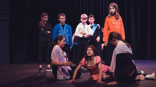 A group of students performing on stage, some sitting on the floor, others crowding around the people sitting