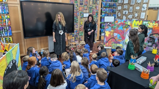 A school hall is decorated with lots of art work and a teacher is standing up, talking to a group of pupils sat on the floor.