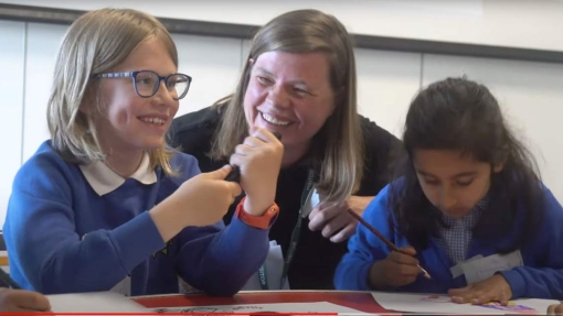 Three children in blue school uniforms are seated at a table, engaged in a creative activity. The child on the left, wearing glasses, holds a black object, possibly a pen or marker. The child in the middle, with long hair, is leaning forward and partially obscured. The child on the right is drawing on paper with a pencil or crayon.