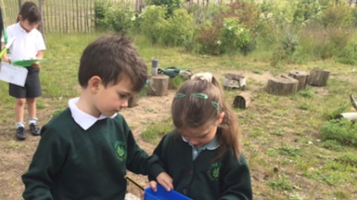 Two children in green school uniforms stand outdoors on grass with tree stumps and greenery in the background. One holds a blue clipboard and magnifying glass; the other holds a blue rectangular container. A third child in the background holds a green clipboard and writes something down.