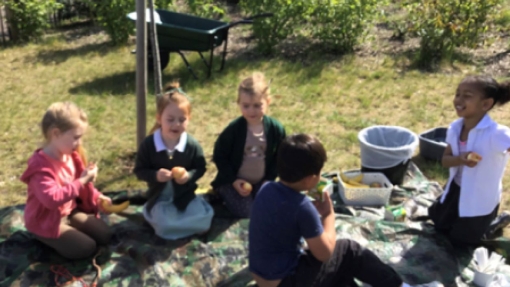 Five children sit on a camouflage-patterned tarp outdoors, engaged in an activity. They wear casual clothing and school uniforms. Around them are water bottles, small containers, two gray bins, a green wheelbarrow, and surrounding greenery.