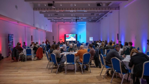 A spacious, modern conference room with high ceilings, exposed beams, and wooden floors. Round tables with blue chairs are arranged throughout the room, each occupied by groups of people engaged in discussions or activities. A large screen at the front displays colorful graphics and text. Blue and pink lighting illuminates the walls, creating a vibrant atmosphere.