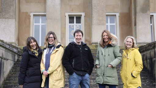 Five people stand on stone steps in front of a large building with columns and windows. They wear winter clothing in colors like black, yellow, and green. The building's facade includes two windows and a central door between the columns.