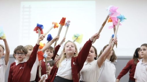Children in school uniforms—white shirts and red sweaters—stand in a classroom holding colorful handmade wands made from sticks and tissue paper. 