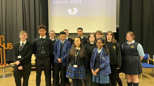 A group of students stand on a stage in front of a screen that reads 'HFCMAT Student Conference March 2024.' They are dressed in various school uniforms, including blazers, ties, and skirts. The stage background features black curtains and several chairs.