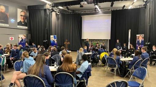 A large room filled with students in school uniforms seated at round tables, engaged in discussions. At the front, a stage with black curtains features a projector screen displaying five discussion prompts related to understanding a topic, its relevance to studies, challenges faced, effective strategies, and helpful resources. Several posters are visible on the walls