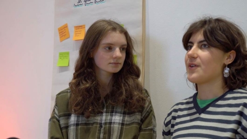 Two individuals with blurred faces stand side by side. The person on the left has long, wavy hair and wears a green plaid shirt. The person on the right has short, curly hair and wears a striped sweater. Behind them is a wall displaying colorful sticky notes and a large sheet of paper with handwritten blue text at the top.