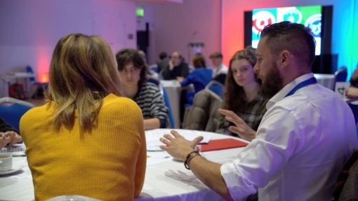 A group of people are seated around a table in a conference room, actively engaged in discussion. In the foreground, a person in a yellow sweater is seen from behind, facing another person who is gesturing with their hands. Additional attendees are visible in the background at other tables, with papers and cups in front of them. A large screen on the right displays colorful graphics.