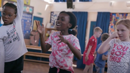 Children in a classroom or gym take part in an activity. One raises an arm, another gestures, and a third wears a 'YOU GOT THIS' shirt. Faces are obscured. Blue curtains and artwork are in the background.