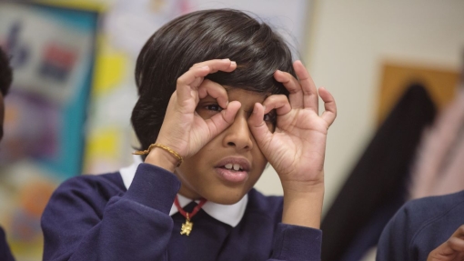 A child in a school uniform, face blurred for privacy, holds up one hand making an 'OK' gesture. The child wears a navy blue sweater with colorful figures and the name 'HANNAH' embroidered on it. In the background, a classroom is visible with blurred posters and another student partially seen on the left