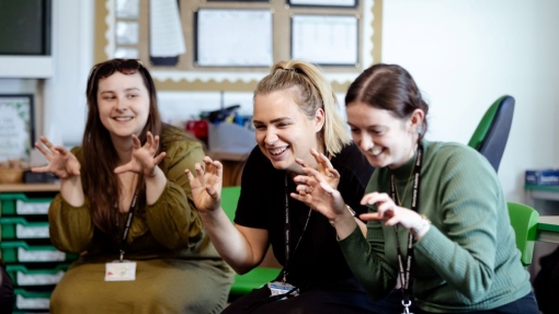 Three individuals seated on green chairs in a classroom, holding their hands up with fingers bent like claws. Their faces are blurred for privacy. Each person wears a lanyard with an ID badge. Behind them, a bulletin board with pinned papers and shelves with various items are visible.