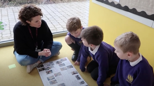 A teacher and three young students sit on the classroom floor, looking at a large poster with images and text. The teacher wears a black sweater and blue jeans; the students are in purple school uniforms. The classroom has yellow walls and a large window revealing an outdoor paved area.