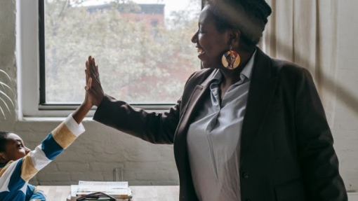 In a classroom, a teacher in a dark blazer and white shirt stands beside a wooden table, high-fiving a student in a striped sweater. The student is seated with notebooks and pencils in front of them. Another student sits at the same table, writing in a notebook. A large window in the background lets in natural light and shows some greenery outside.