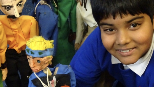 A person wearing a blue sweater with a white collar is holding a marionette puppet with yellow hair, dressed in blue clothing with black and orange patches. In the background, several other marionette puppets are visible, including one in an orange shirt and another in green pants. All faces, including the person's and the puppets