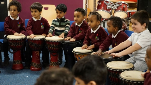 Young children in school uniforms sit in a semi-circle, each playing a drum. One child wears a green and white striped sweater. A set of drums is visible on a stage in the background.