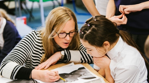 In a classroom, a teacher and student sit at a table working on an art project. The teacher paints on a black-and-white image of an animal, possibly a dog, while the student watches. Other students stand nearby observing. Newspapers cover the table to protect it from paint.