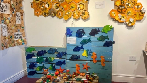 Gallery exhibit with marine-themed art on blue panels, honeycomb artworks with bees above, a quilt-like piece on the left wall, and recycled material sculptures on a wooden pallet.