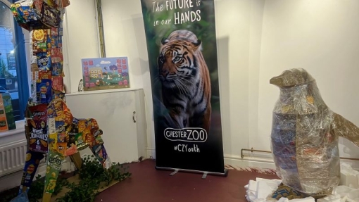 A giraffe sculpture made from snack wrappers, a tiger-themed banner, and a penguin sculpture wrapped in plastic are displayed in a room with white walls and a maroon floor.