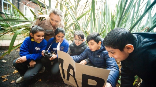 A group of children and an adult outdoors among tall plants, with one child holding a cardboard sign labeled 'MAP' and another holding a camera. A building and greenery are visible in the background.