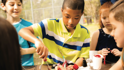 A group of children gathered around an outdoor table, working on an electronics project. The central child, in a yellow and blue striped shirt, holds wires and tools, while others also handle electronic components. A fence and trees are visible in the background.